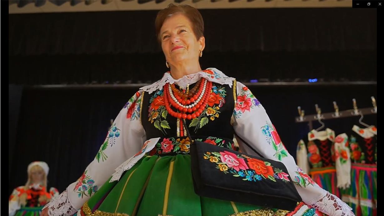 Kobieta ubrana w tradycyjny strój ludowy, składający się z białej bluzki z kolorowymi kwiatowymi haftami, czarnego gorsetu z kwiatowymi wzorami, zielonej spódnicy oraz czerwonych korali. Na ramieniu ma czarną torebkę z kwiatowym wzorem. W tle widać inne stroje ludowe na wieszakach.