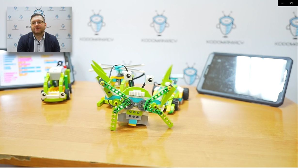 Na zdjęciu widać stół z trzema robotami z klocków LEGO w różnych kolorach, głównie zielonym i żółtym. W tle znajduje się tablet z jasnym ekranem oraz plakat z logo "KODOMANIACY".