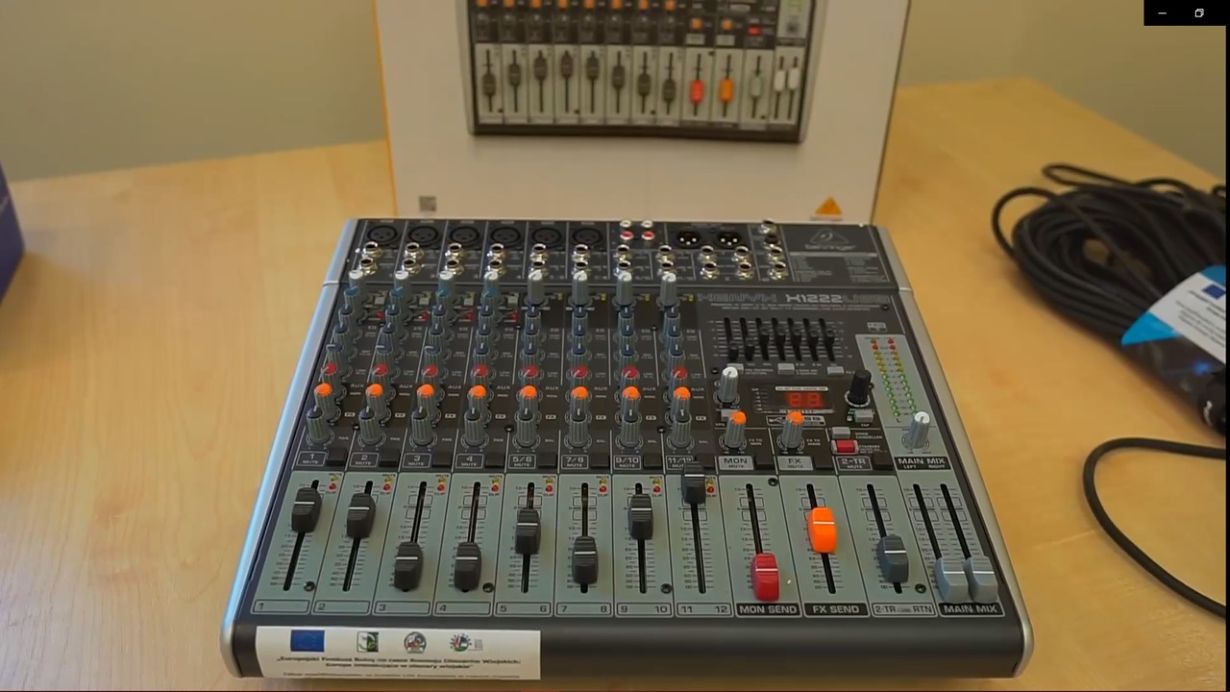 Na zdjęciu znajduje się mikser audio umieszczony na drewnianym stole. Mikser ma wiele suwaków, pokręteł i przycisków, które służą do regulacji dźwięku. W tle widać pudełko z obrazem tego samego miksera oraz zwinięty kabel po prawej stronie. Na mikserze znajduje się naklejka informująca o finansowaniu projektu z UE.