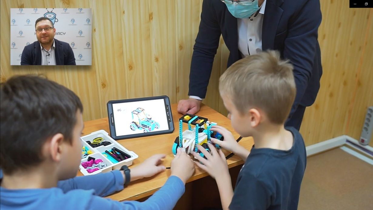 Dwóch chłopców siedzi przy stole, budując robota z klocków LEGO. Na stole znajduje się tablet z instrukcją budowy robota oraz pudełko z częściami. Obok nich stoi dorosły mężczyzna w garniturze i maseczce, obserwując ich pracę.