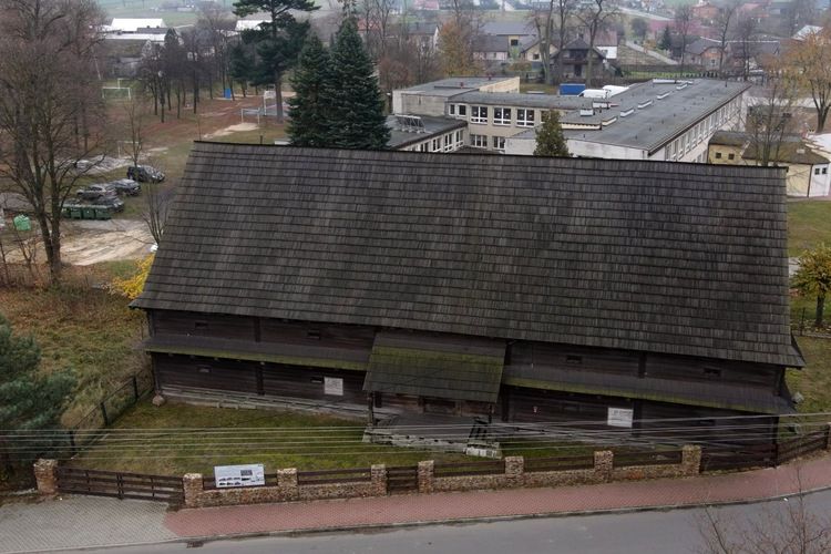 Zdjęcie przedstawia drewniany budynek z dużym, spadzistym dachem, otoczony ogrodzeniem z cegły. W tle widać nowoczesne budynki i drzewa. Budynek może być zabytkowy, co czyni go interesującym w kontekście architektury i historii.