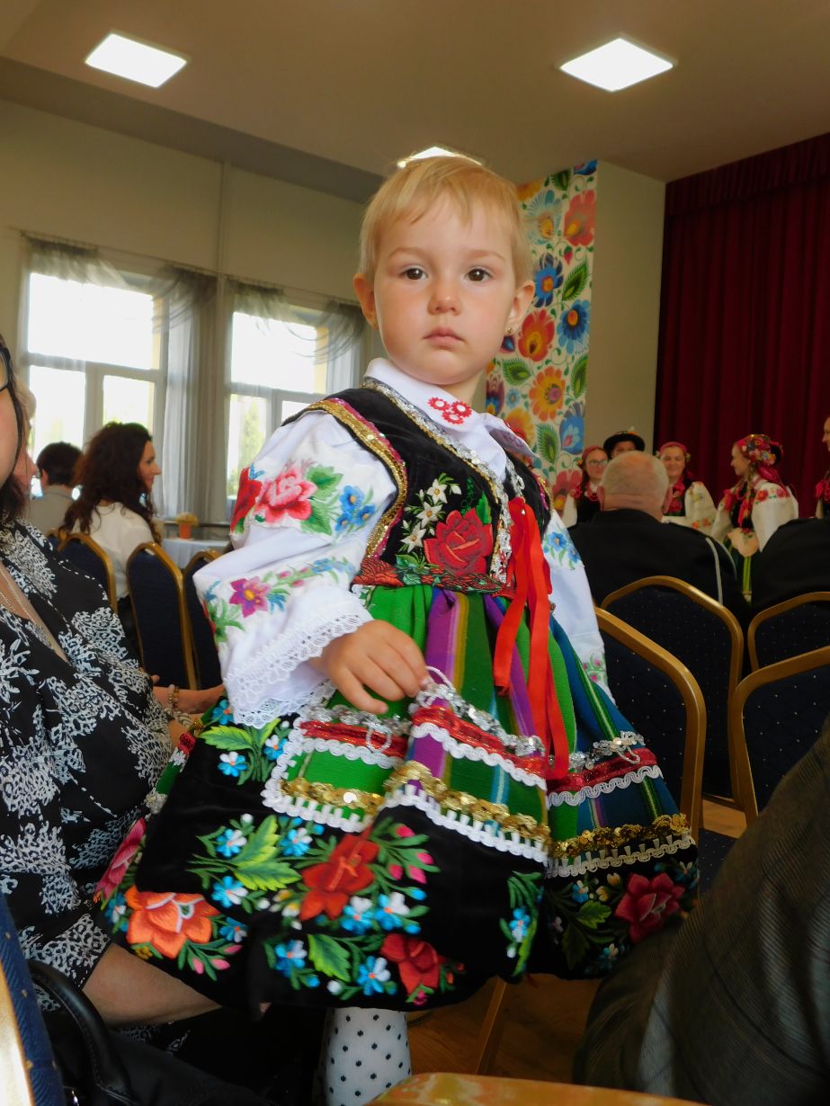 Dziecko w tradycyjnym stroju ludowym siedzi na kolanach osoby dorosłej. Strój jest kolorowy, z dominującymi kolorami zielonym, czarnym i czerwonym, ozdobiony kwiatowymi haftami i koronkami. W tle widać innych ludzi w podobnych strojach oraz kolorową dekorację ścienną.