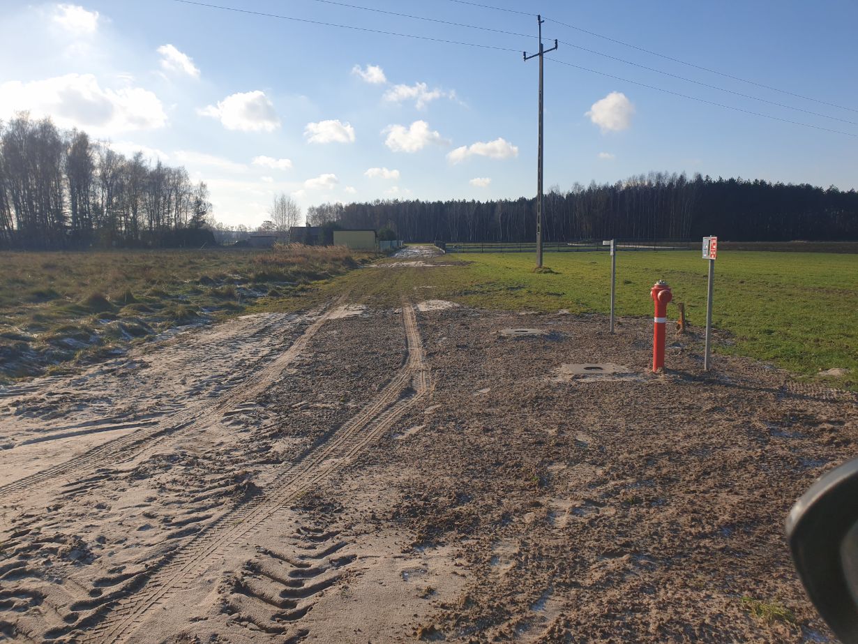 Zdjęcie przedstawia wiejską drogę gruntową prowadzącą przez otwarte pole z trawą i drzewami w tle. Na pierwszym planie po prawej stronie znajduje się czerwony hydrant przeciwpożarowy oraz słup energetyczny z przewodami. W tle widać las i kilka budynków.