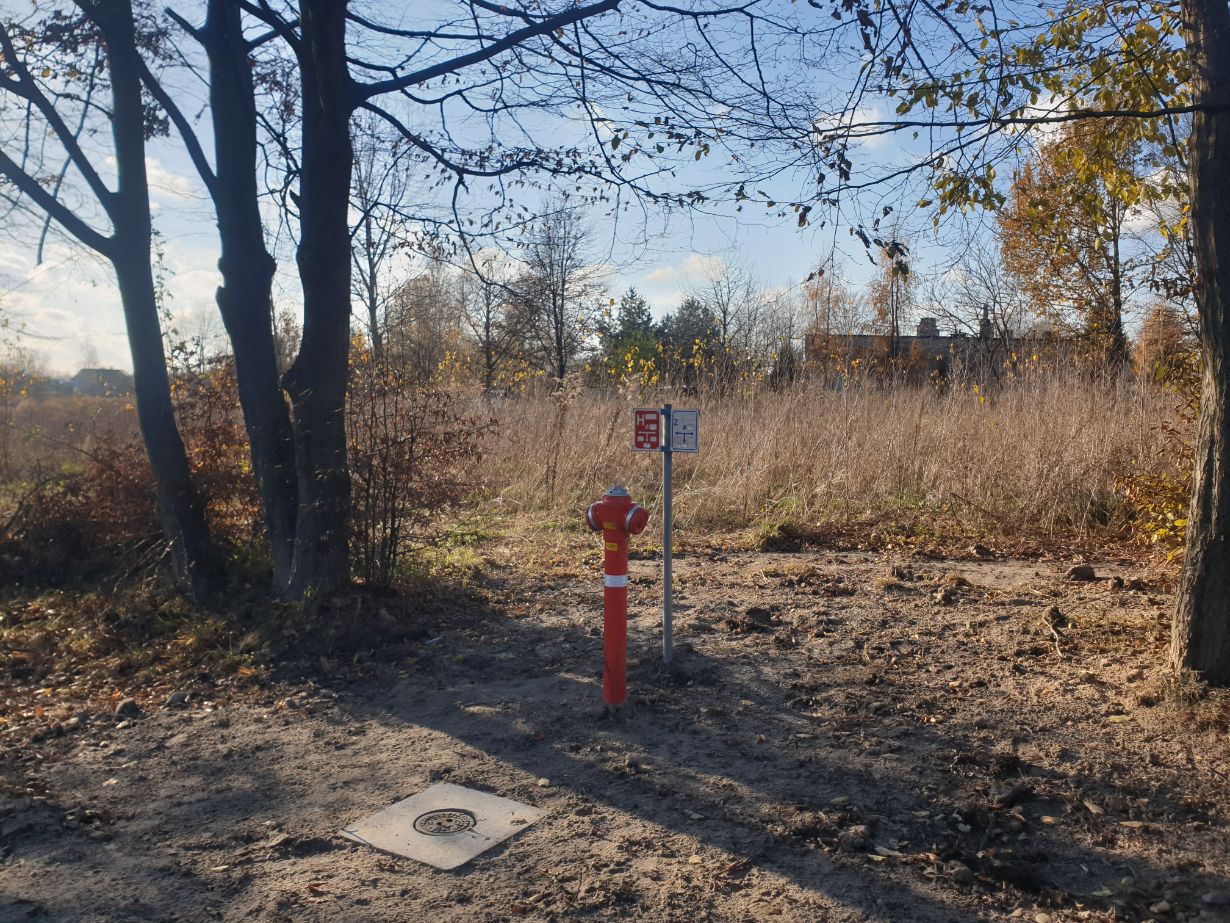 Zdjęcie przedstawia hydrant przeciwpożarowy na skraju zarośniętego terenu, otoczony drzewami bez liści. Obok hydrantu znajduje się znak informacyjny. W tle widać budynki i niebo z chmurami.