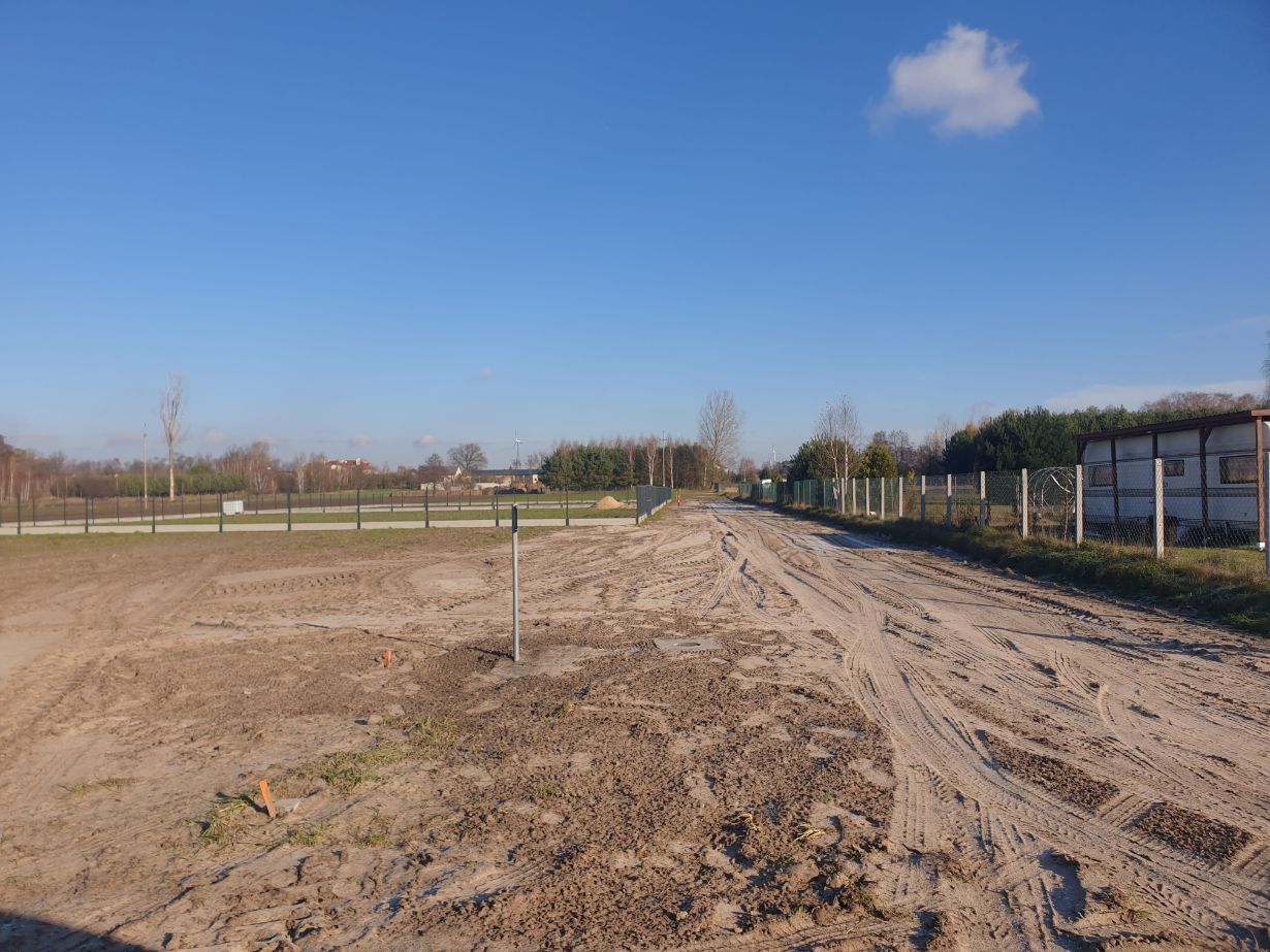 Zdjęcie przedstawia plac budowy z piaszczystą drogą i ogrodzeniem po prawej stronie. W tle widać drzewa i niebieskie niebo z kilkoma chmurami. Na pierwszym planie znajduje się metalowy słupek wbity w ziemię.