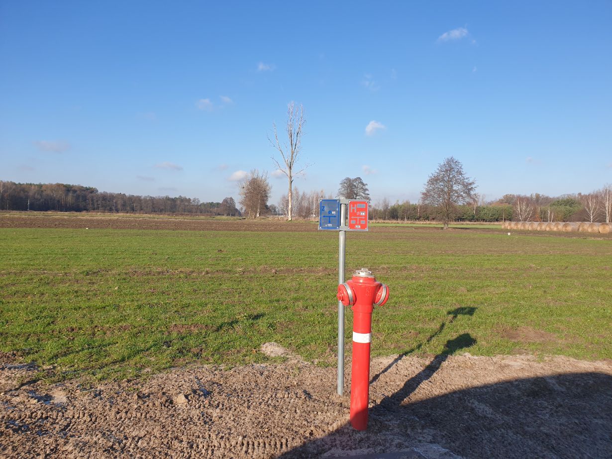 Zdjęcie przedstawia hydrant przeciwpożarowy na tle otwartego pola z kilkoma drzewami w oddali. Hydrant jest czerwony z białym paskiem, a obok niego znajduje się metalowy słupek z dwoma znakami informacyjnymi: niebieskim i czerwonym.