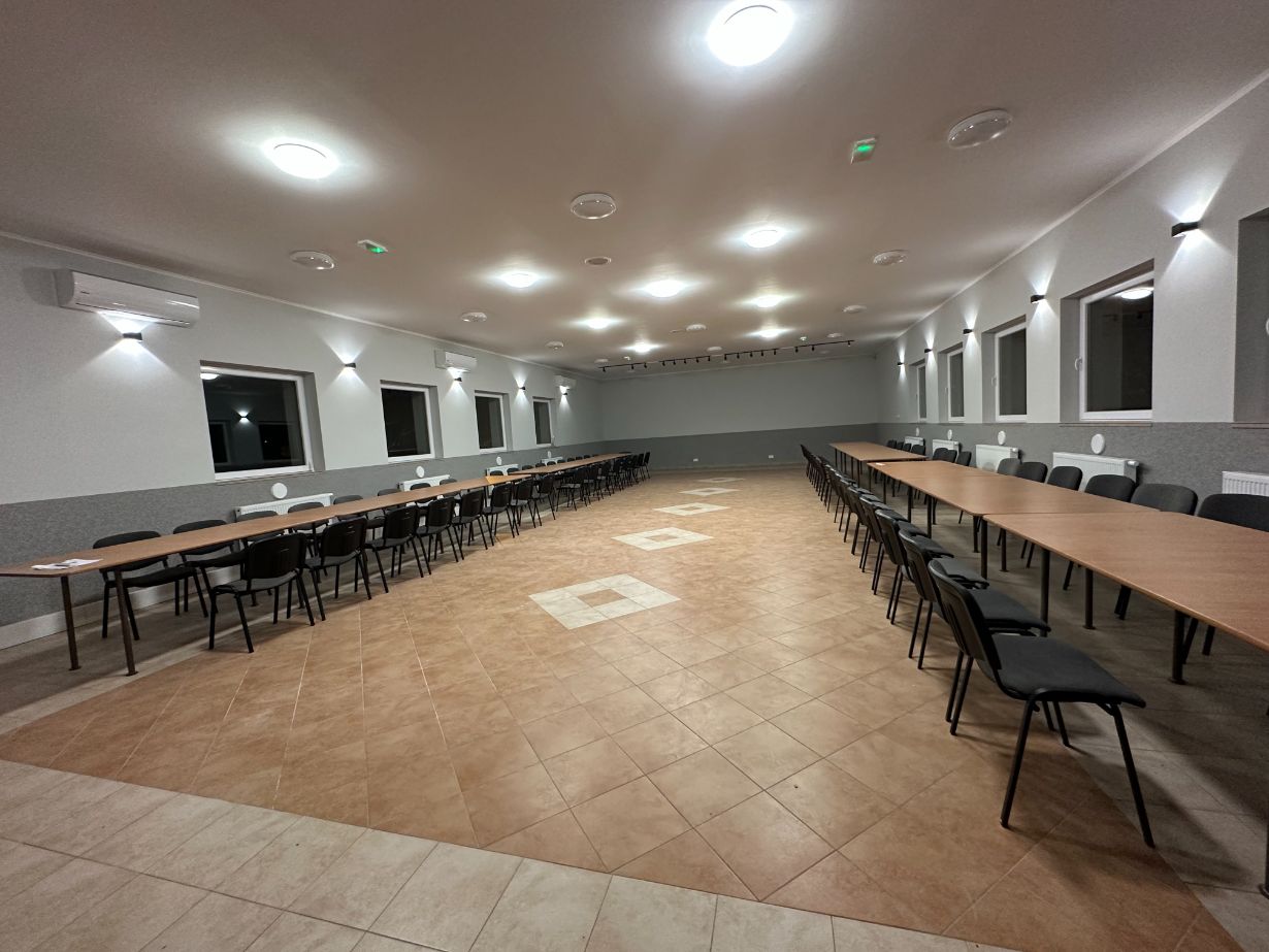 Zdjęcie przedstawia dużą, nowoczesną salę konferencyjną z długimi stołami ustawionymi w kształt litery "U". Wzdłuż stołów ustawione są krzesła. Sala jest dobrze oświetlona, z licznymi lampami sufitowymi i kinkietami na ścianach. Na ścianach znajdują się okna, a na suficie zamontowane są klimatyzatory.