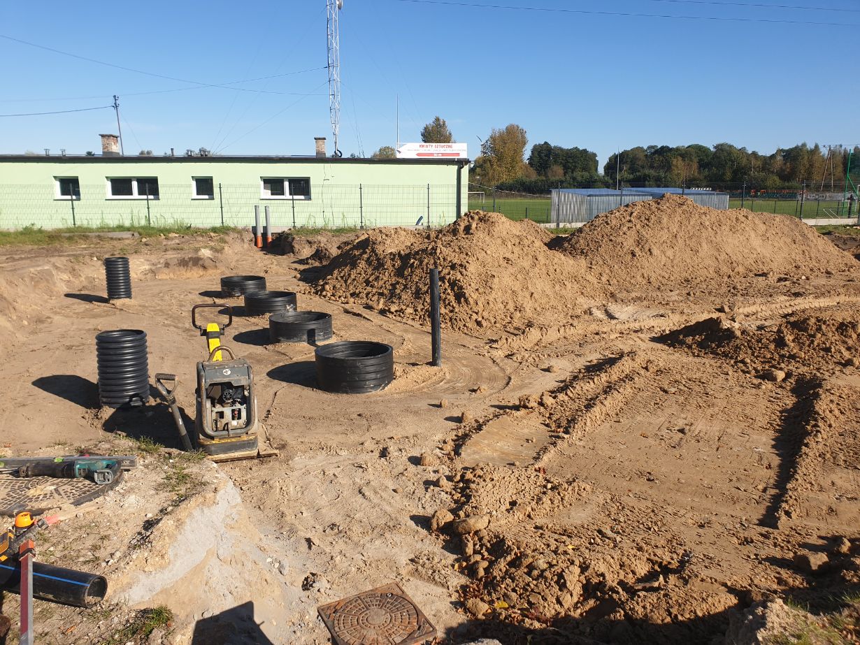 Zdjęcie przedstawia plac budowy z wykopami i rurami kanalizacyjnymi. W tle widać zielony budynek oraz kopce ziemi. Na pierwszym planie znajduje się zagęszczarka do gruntu.