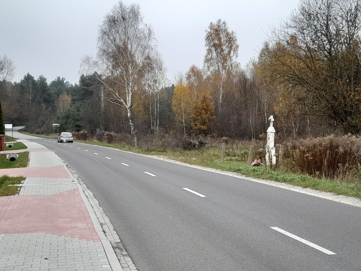 Zdjęcie przedstawia asfaltową drogę biegnącą przez wiejską okolicę, z chodnikiem po lewej stronie. Po prawej stronie drogi znajduje się przydrożny krzyż z kwiatami u podstawy. W tle widać drzewa w jesiennych kolorach.