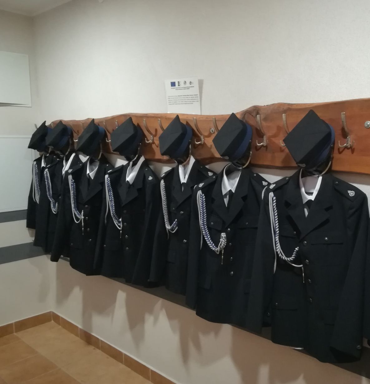 Na zdjęciu widać sześć mundurów strażackich wiszących na drewnianym wieszaku na ścianie. Każdy mundur składa się z czarnej marynarki, białej koszuli, krawata oraz czapki. Na wieszaku znajduje się również tabliczka informacyjna.