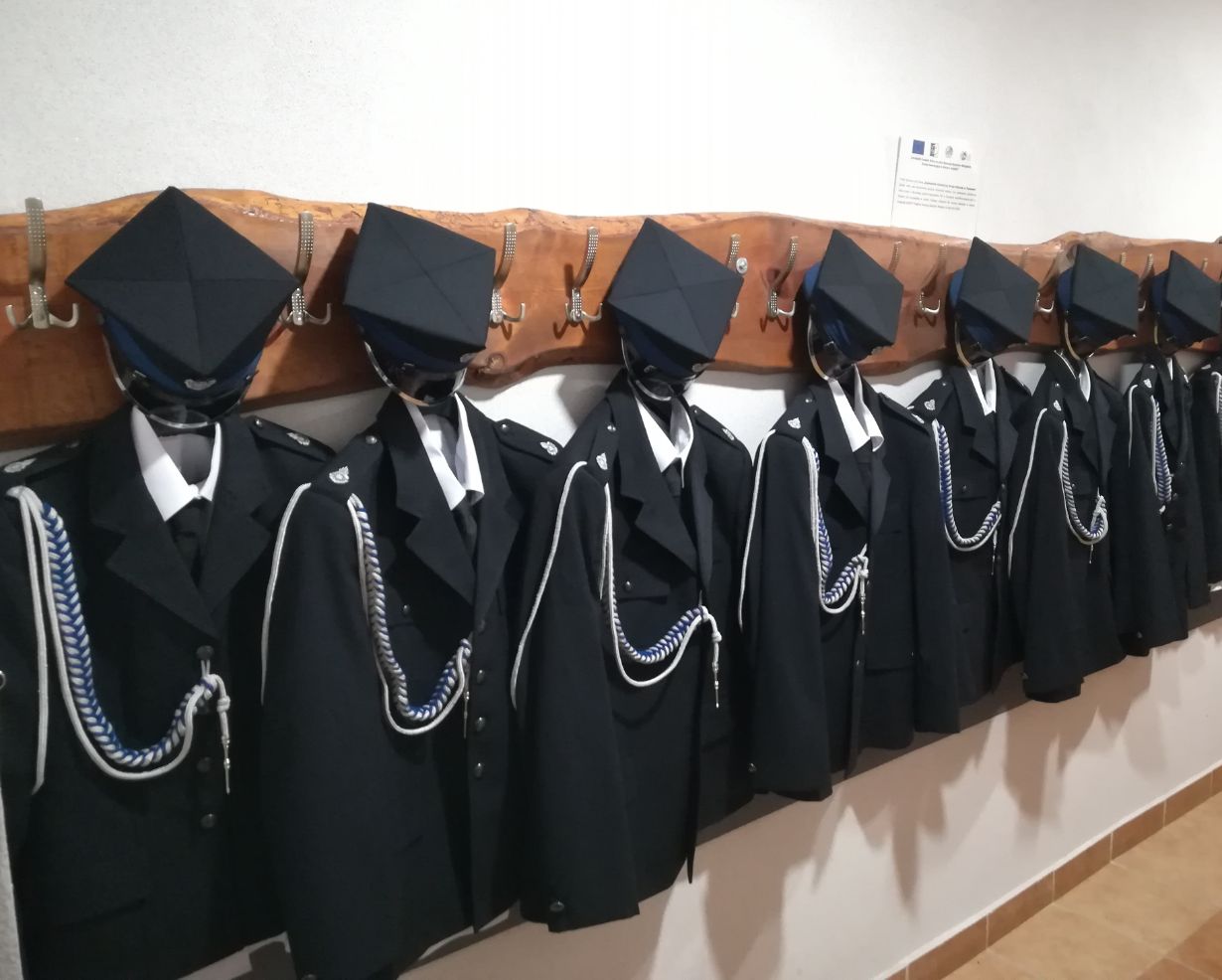 Na zdjęciu widać rząd mundurów strażackich wiszących na drewnianym wieszaku. Każdy mundur składa się z czarnej marynarki z białymi i niebieskimi sznurami oraz czapki. W tle znajduje się tablica informacyjna.