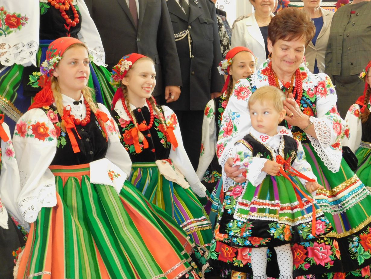 Zdjęcie przedstawia grupę ludzi ubranych w tradycyjne stroje ludowe, głównie kobiety i dzieci, z kolorowymi spódnicami, haftowanymi bluzkami i koralami. Stroje mają motywy kwiatowe i paski w żywych kolorach, takich jak zielony, czerwony i biały.