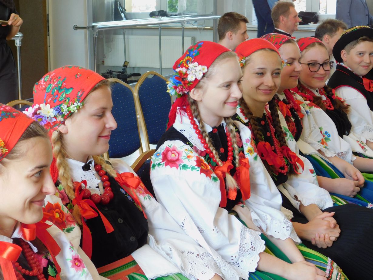 Zdjęcie przedstawia grupę ludzi ubranych w tradycyjne stroje ludowe, siedzących w rzędzie. Stroje obejmują kolorowe chusty z kwiatami, białe bluzki z haftem, czarne kamizelki i pasiaki. Osoby trzymają się za ręce, co sugeruje jedność lub udział w wydarzeniu kulturalnym.