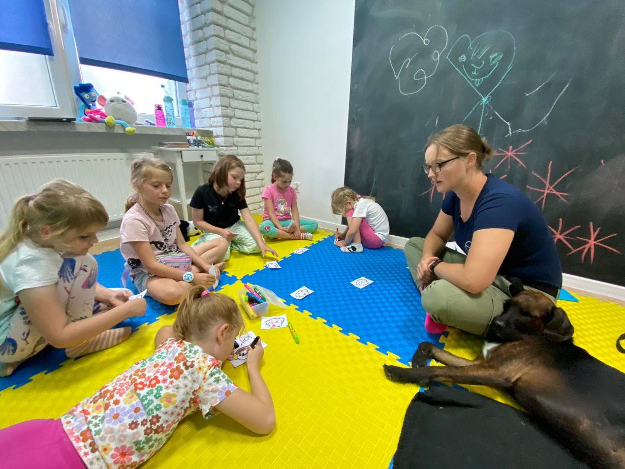 Na zdjęciu widać grupę dzieci siedzących na kolorowej macie w pokoju z dużą tablicą kredową. Dzieci rysują i kolorują obrazki, a obok nich siedzi dorosła osoba. Na tablicy kredowej są narysowane serca i postacie. Obok dorosłej osoby leży pies.