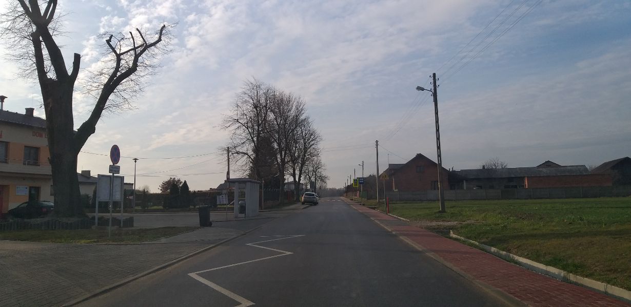 Zdjęcie przedstawia wiejską drogę z chodnikiem po prawej stronie. Po lewej stronie znajduje się budynek z napisem "DOM", przystanek autobusowy i znak drogowy. W tle widać kilka domów i drzewa bez liści.