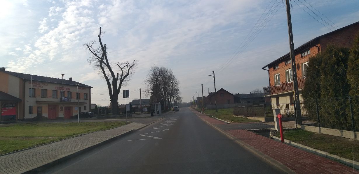 Zdjęcie przedstawia ulicę w małym miasteczku. Po lewej stronie znajduje się budynek z napisem "OCHOTNICZA STRAŻ POŻARNA CHORZĘCIN". Po prawej stronie widać domy mieszkalne i drzewa. Na ulicy zaparkowany jest samochód.