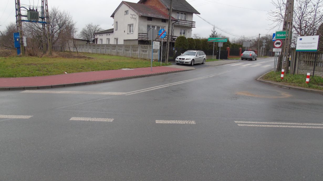 Na zdjęciu widać skrzyżowanie dróg z oznakowaniem poziomym i pionowym. Po lewej stronie znajduje się trawnik z niebieską skrzynką elektryczną, a po prawej dom i zaparkowany samochód. W tle widoczne są znaki drogowe i inne pojazdy.