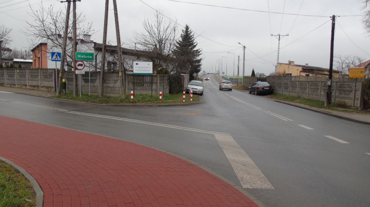 Zdjęcie przedstawia skrzyżowanie dróg w miejscowości Niebórz. Widać znaki drogowe, w tym znak zakazu skrętu w lewo oraz znak przejścia dla pieszych. Na drodze znajdują się samochody, a w tle widoczny jest most.