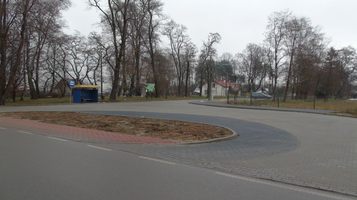 Zdjęcie przedstawia pusty przystanek autobusowy na tle parku z drzewami bez liści. W tle widać również budynek z czerwonym dachem oraz zaparkowany samochód. Droga jest wyłożona kostką brukową, a na pierwszym planie znajduje się zakręt.