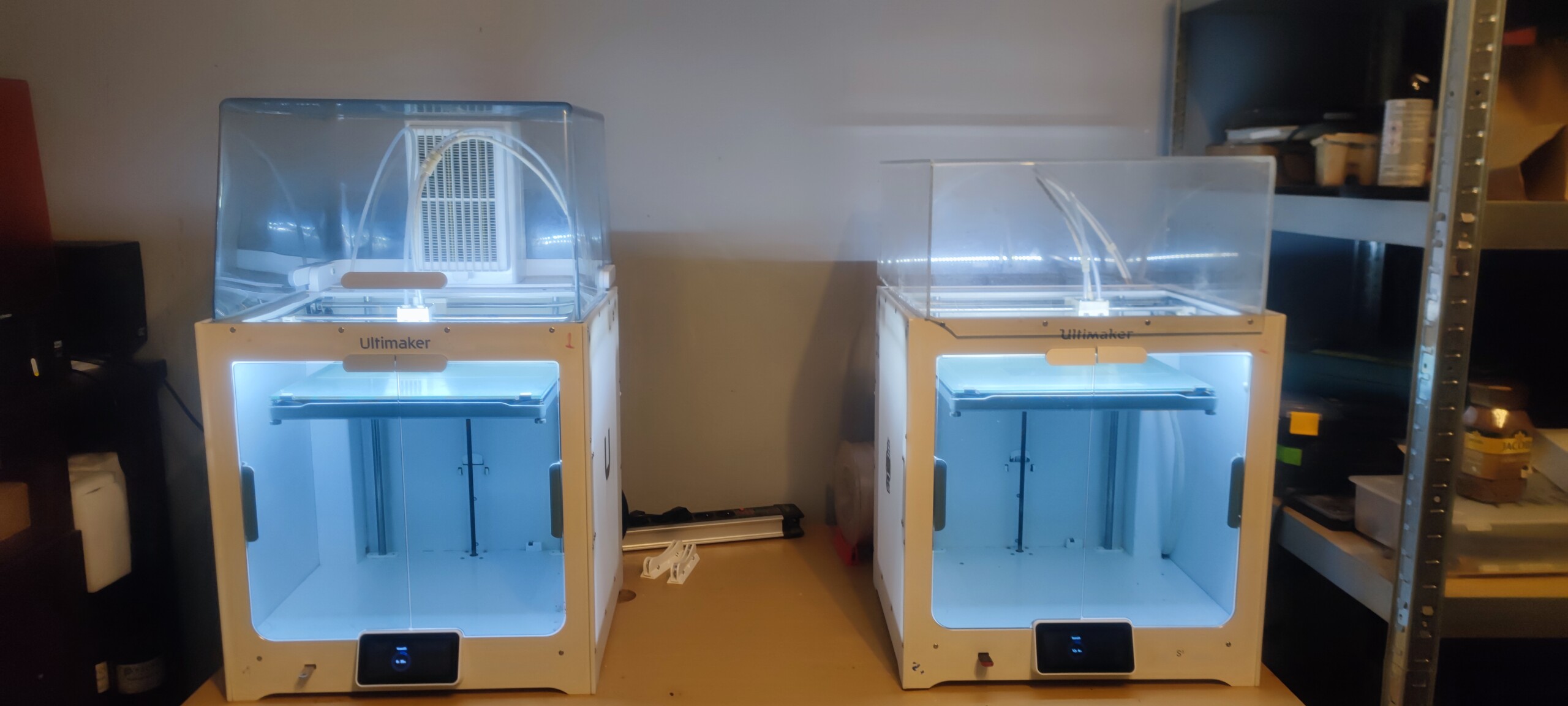 Obraz przedstawia dwie drukarki 3D marki Ultimaker ustawione obok siebie na stole. Obie drukarki mają przezroczyste osłony i są podświetlone wewnętrznym niebieskim światłem.