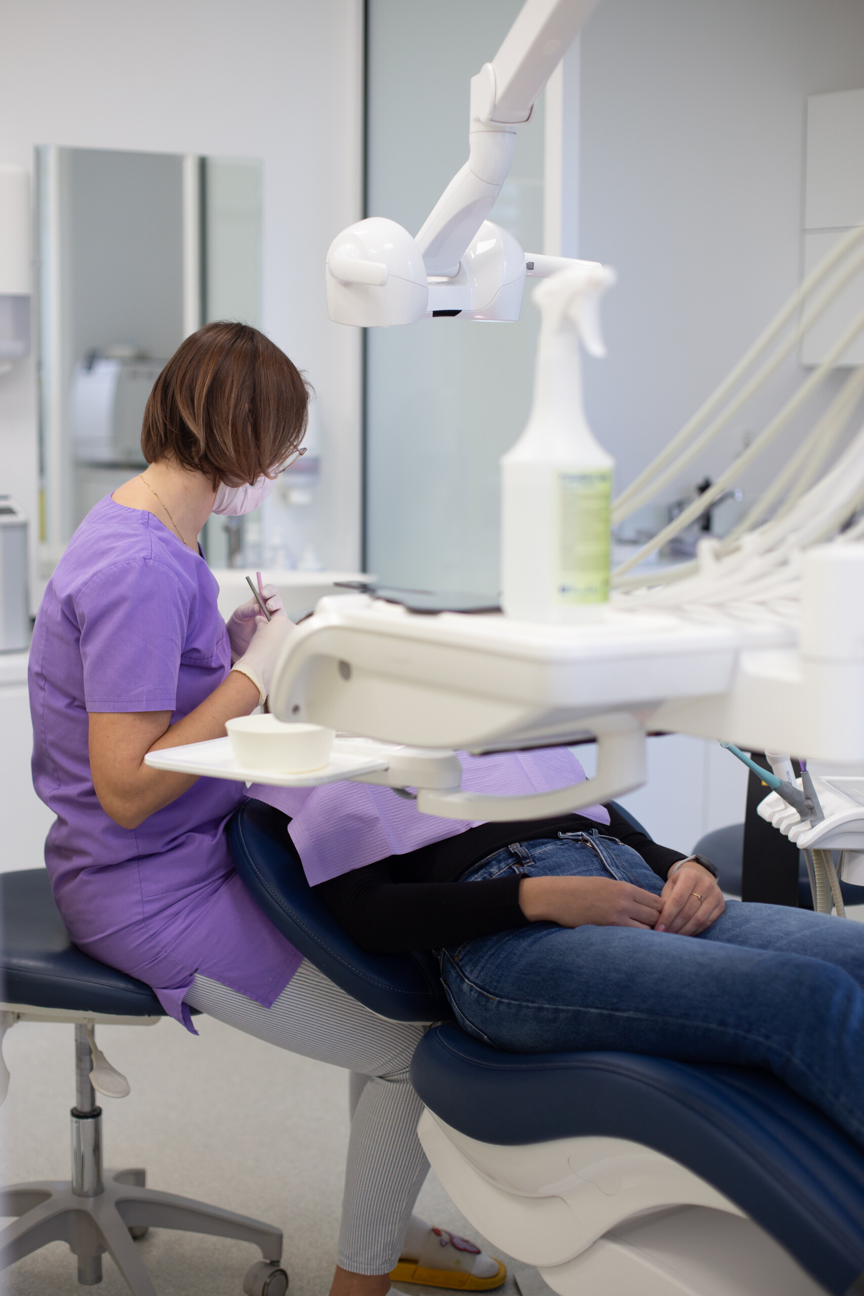 Dentysta w fioletowym uniformie i maseczce zajmuje się pacjentem leżącym na fotelu. Nad pacjentem znajduje się lampa dentystyczna, a w tle widoczne są różne sprzęty i materiały dentystyczne.