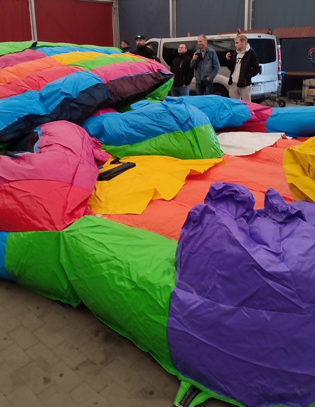 Na zdjęciu widać dużego, kolorowego dmuchańca, który jest złożony i leży na ziemi w zamkniętym pomieszczeniu. W tle znajduje się kilka osób stojących obok białego vana. Kolory dmuchańca to m.in.: zielony, niebieski, różowy, fioletowy, żółty i czerwony.