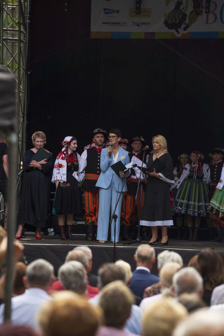 Na zdjęciu widać kobietę w błękitnym garniturze przemawiającą na scenie. W tle znajduje się baner z napisem "50 lat REGIONALNEGO ZESPOŁU PIEŚNI I TAŃCA BOCZKI CHEŁMOŃSKIE". Pozostali ludzie są ubrani w tradycyjne stroje ludowe.