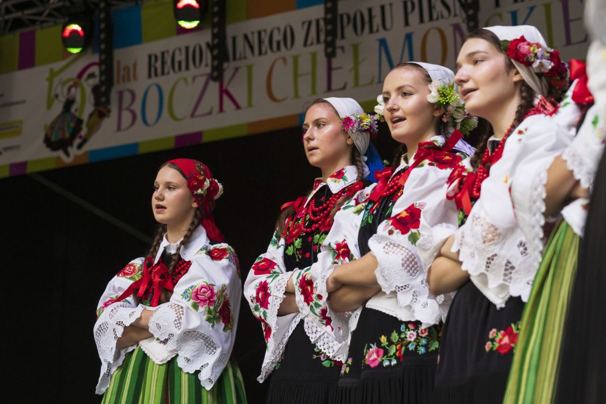 Na zdjęciu widać grupę osób ubranych w tradycyjne stroje ludowe, stojących na scenie. W tle znajduje się baner z napisem "50 lat REGIONALNEGO ZESPOŁU PIEŚNI I TAŃCA BOCZKI CHEŁMOŃSKIE".