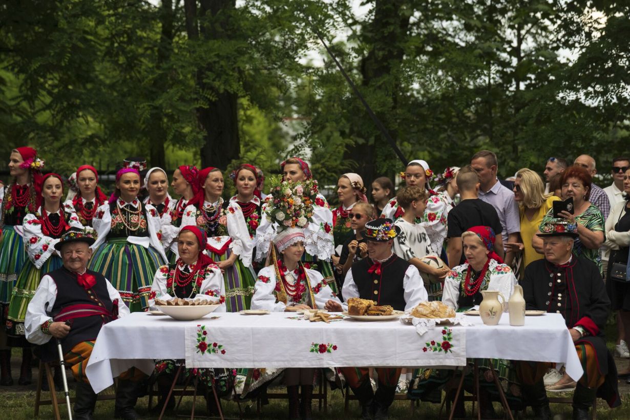 Grupa ludzi ubrana w tradycyjne stroje ludowe zgromadzona na świeżym powietrzu. Kobiety mają kolorowe spódnice, haftowane bluzki i korale, a mężczyźni noszą kamizelki i kapelusze. Na stole z przodu, przykrytym białym obrusem z kwiatowym haftem, znajdują się różne potrawy i napoje. W tle widać zielone drzewa sugerujące, że wydarzenie odbywa się w parku lub ogrodzie.