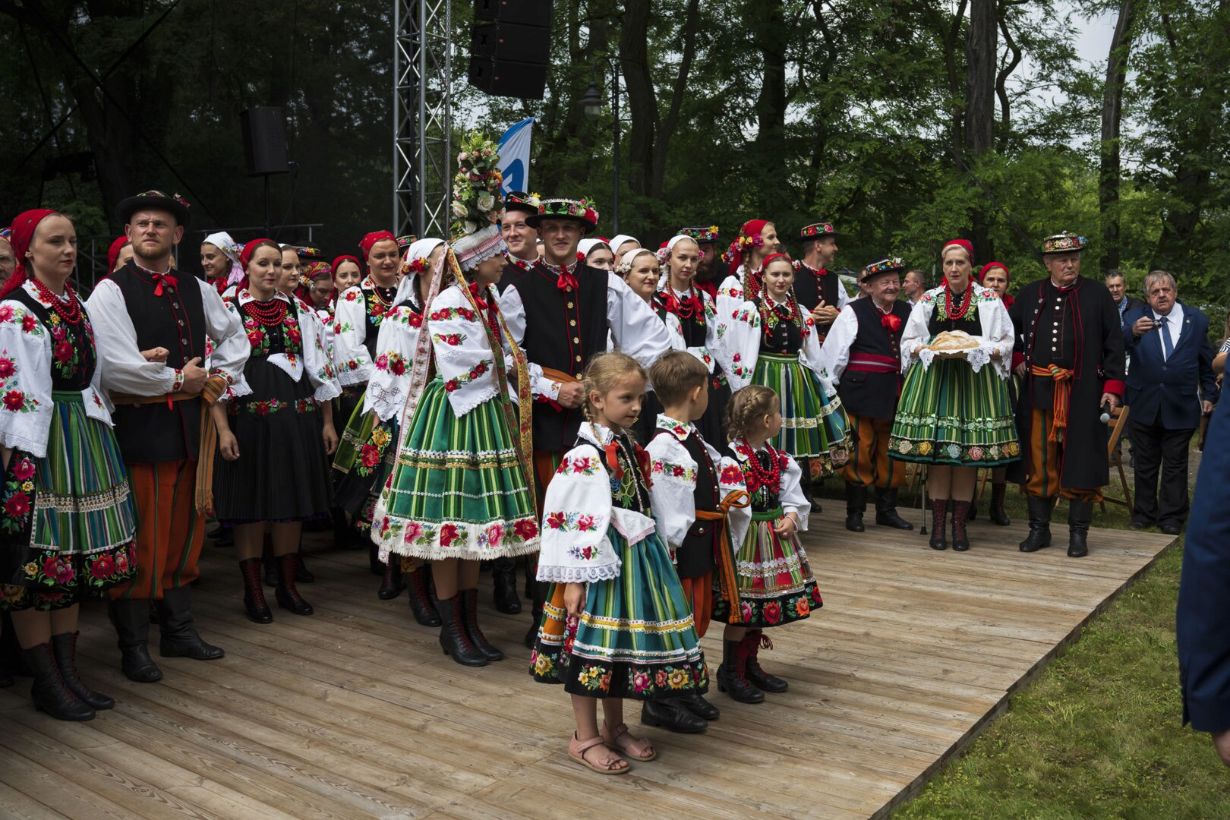 Grupa ludzi ubrana w tradycyjne stroje ludowe stoi na drewnianej scenie na tle drzew. Kobiety i dzieci mają na sobie zielone spódnice z kwiatowymi wzorami, białe bluzki i czerwone korale, a mężczyźni noszą czarne kamizelki i pomarańczowe spodnie.