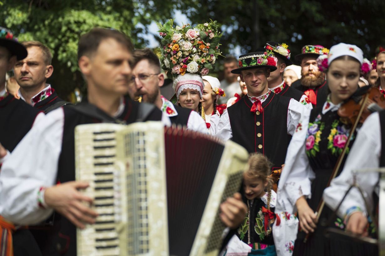 Grupa ludzi w tradycyjnych strojach ludowych bierze udział w wydarzeniu kulturalnym. Ubrani są w haftowane stroje z kwiatowymi wzorami, a niektórzy grają na instrumentach muzycznych, takich jak akordeon i skrzypce. Jedna osoba trzyma duży bukiet kwiatów na głowie.