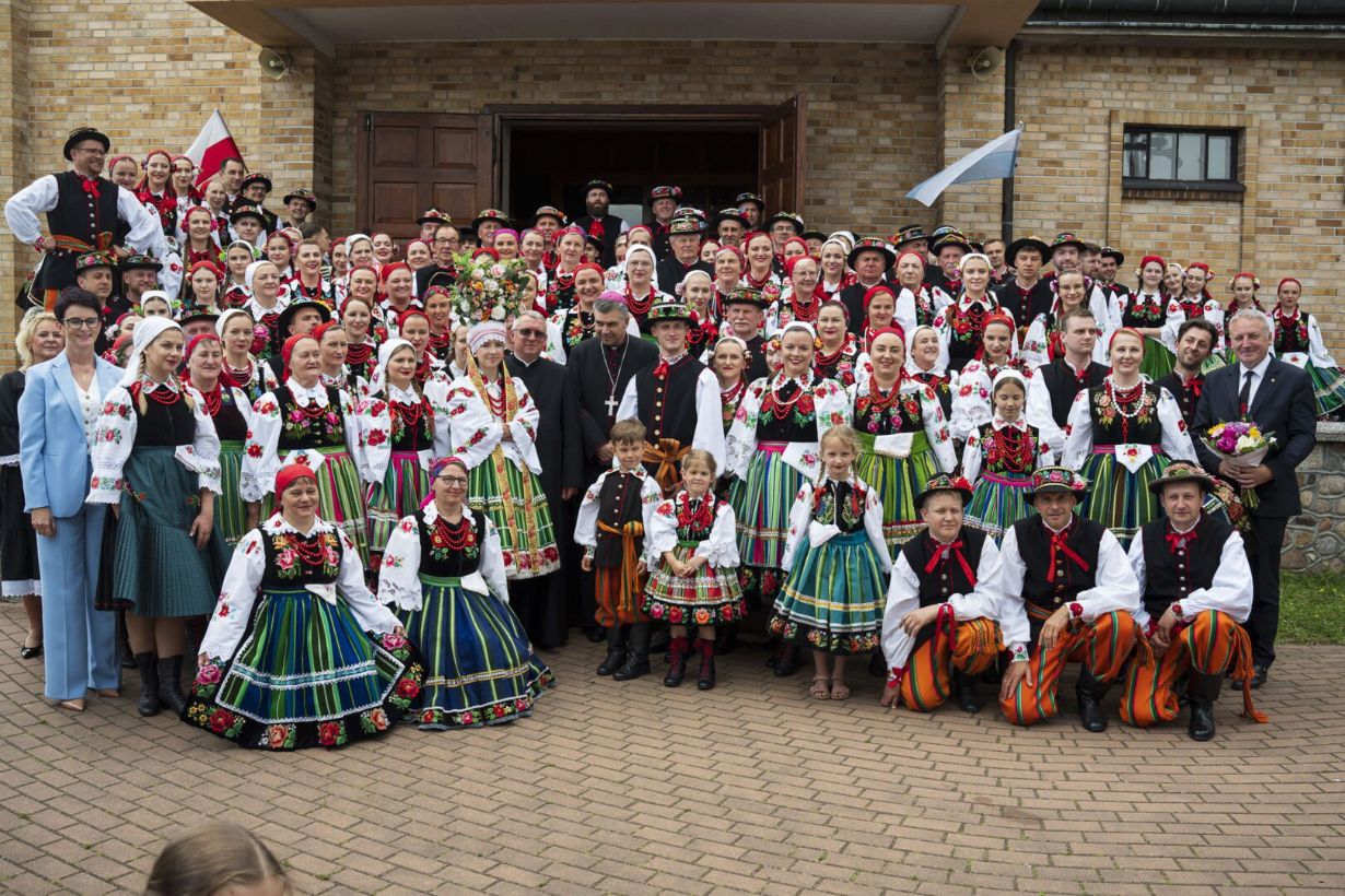 Grupa ludzi w tradycyjnych strojach ludowych pozuje do zdjęcia przed budynkiem. Kobiety mają kolorowe spódnice z kwiatowymi wzorami, białe bluzki i korale, a mężczyźni noszą haftowane kamizelki i pasiaste spodnie. Wygląda na to, że to wydarzenie kulturalne lub świąteczne.