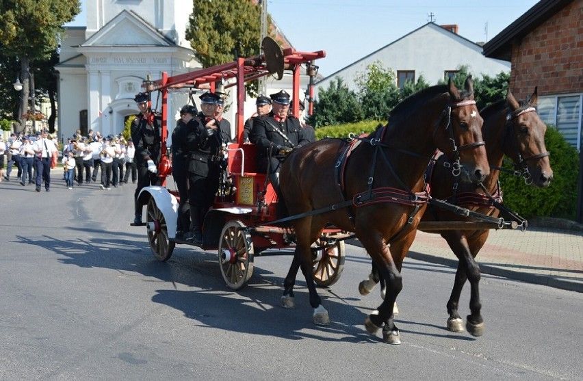 Na zdjęciu widać konny wóz strażacki z dwoma końmi, prowadzony przez strażaków w czarnych mundurach. W tle znajduje się grupa ludzi maszerujących w paradzie oraz budynki mieszkalne i kościół.