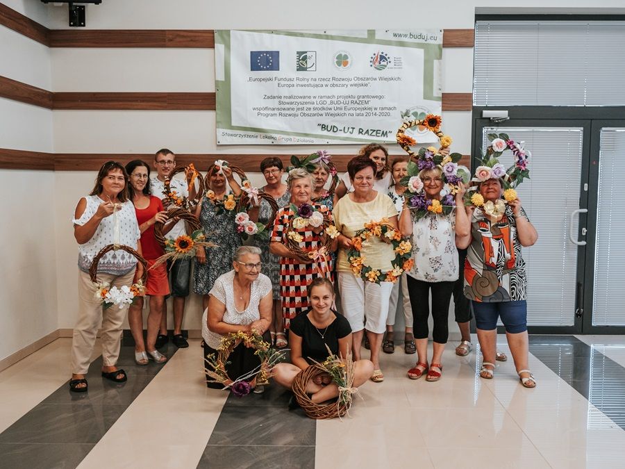 Grupa ludzi trzymających wieńce kwiatowe pozuje do zdjęcia w pomieszczeniu. W tle znajduje się baner z informacjami o projekcie "BUD-UJ RAZEM" współfinansowanym przez Unię Europejską w ramach Programu Rozwoju Obszarów Wiejskich na lata 2014-2020.
