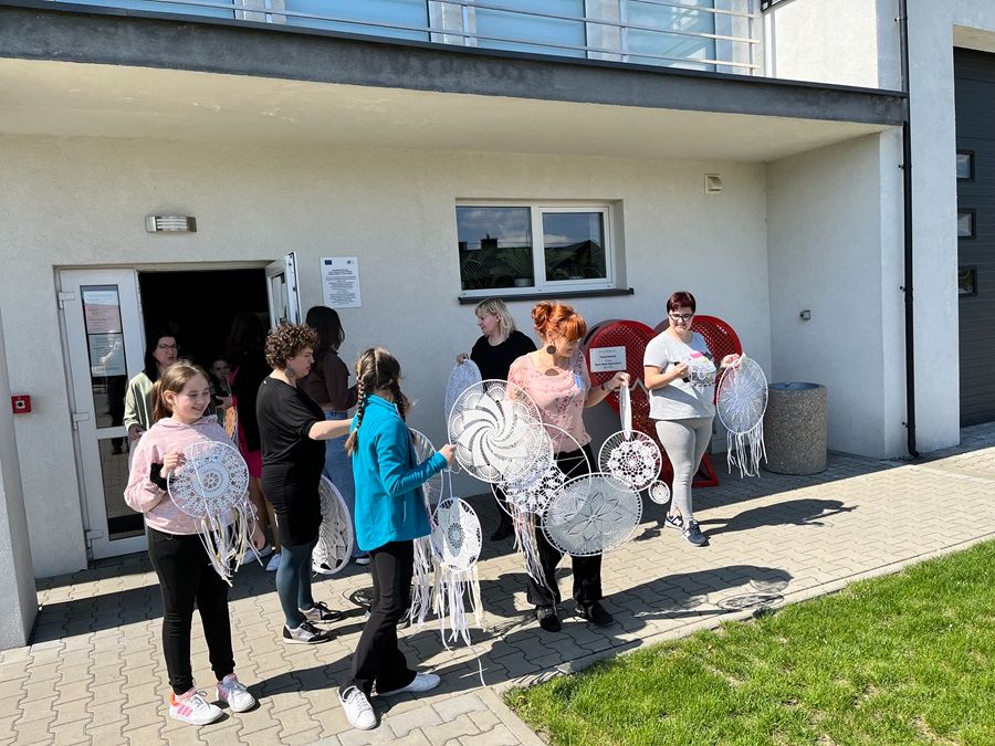 Grupa ludzi stoi na zewnątrz budynku, trzymając duże, ozdobne łapacze snów. W tle widać otwarte drzwi, okno i czerwone krzesła. Scena sugeruje wspólne zajęcia lub warsztaty artystyczne.