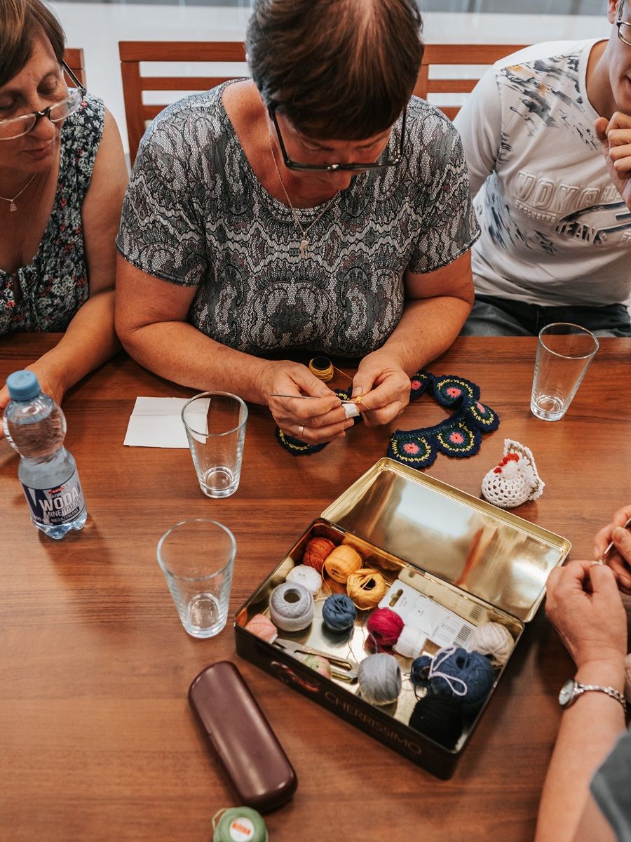 Na zdjęciu widać grupę osób siedzących przy stole, zajmujących się robótkami ręcznymi. Na stole znajduje się metalowe pudełko z różnokolorowymi nićmi, kilka szklanek, butelka wody oraz etui na okulary.