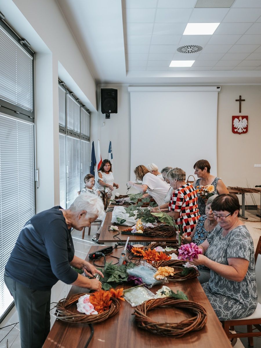 Grupa starszych osób siedzi przy stole, zajmując się rękodziełem. Tworzą ozdobne wieńce z różnych sztucznych kwiatów i liści. Na stole leżą materiały do rękodzieła, takie jak nożyczki, wstążki i drut florystyczny. Scena sugeruje wspólne zajęcia w warsztatach.