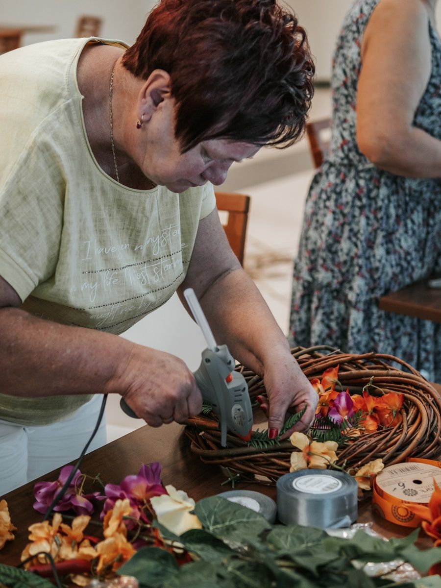 Osoba w żółtej koszulce używa pistoletu do kleju na gorąco, aby przymocować sztuczne kwiaty do wiklinowego wieńca. Na stole znajdują się różne materiały do rękodzieła, w tym taśma klejąca, sztuczne liście i kwiaty.