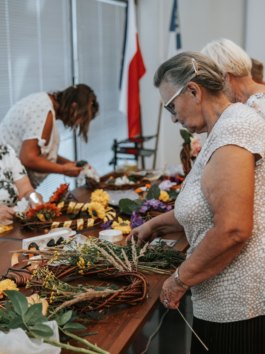 Kilka osób bierze udział w warsztatach florystycznych, tworząc kompozycje kwiatowe na stole. Na stole znajdują się różne materiały florystyczne, takie jak kwiaty, liście, wstążki i narzędzia. W tle widoczne są flagi i okno z żaluzjami.