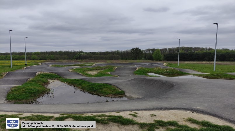 Zdjęcie przedstawia pumptrack w Andrespolu. Na pierwszym planie widoczne są asfaltowe ścieżki z licznymi wzniesieniami i zakrętami, otoczone trawą. W tle znajdują się latarnie oraz drzewa na horyzoncie pod pochmurnym niebem.