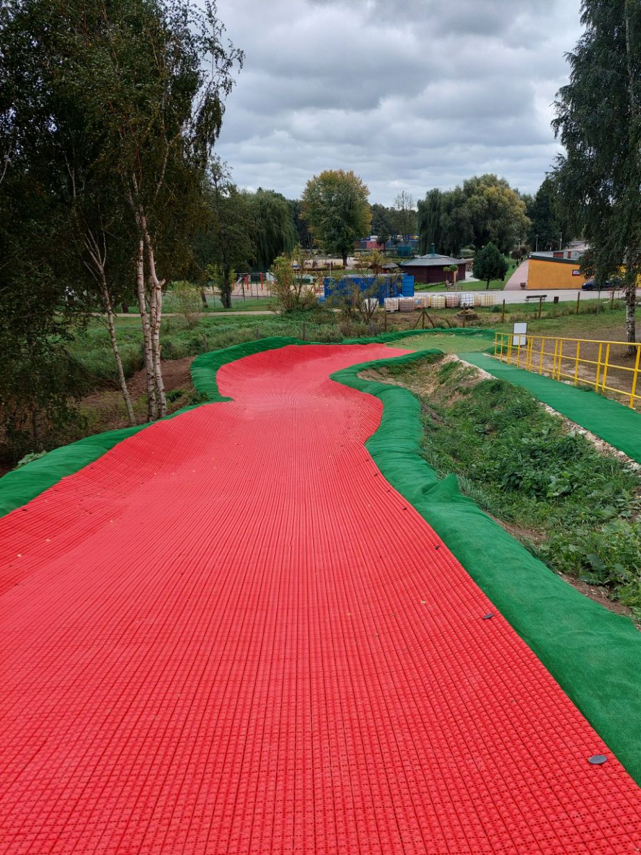 Na zdjęciu widać zakręconą ścieżkę pokrytą czerwonym materiałem, otoczoną zieloną trawą i drzewami. W tle znajduje się park z drzewami, budynkami i placem zabaw. Na niebie są chmury, co sugeruje pochmurny dzień.