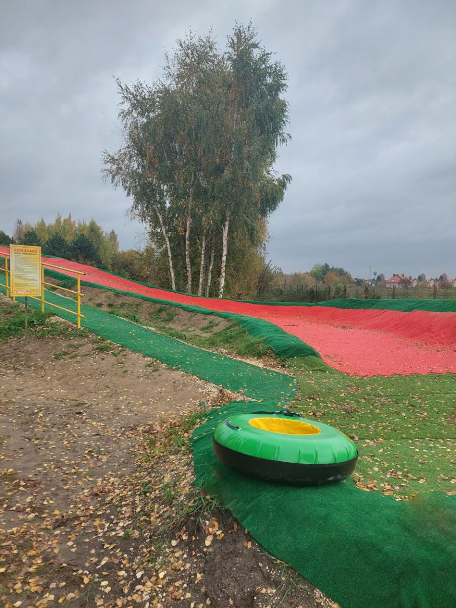 Na zdjęciu widać tor do zjeżdżania na pontonach, pokryty zieloną i czerwoną matą. Na pierwszym planie znajduje się zielony ponton. W tle widać drzewa i tablicę z regulaminem.