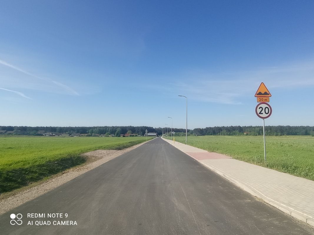 Zdjęcie przedstawia prostą asfaltową drogę biegnącą przez wiejską okolicę, otoczoną zielonymi polami. Po prawej stronie drogi znajduje się znak drogowy ostrzegający o nierównościach na drodze za 30 metrów oraz ograniczenie prędkości do 20 km/h