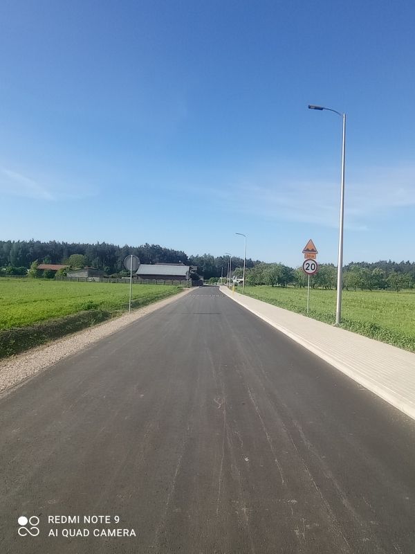 Zdjęcie przedstawia asfaltową drogę biegnącą przez wiejską okolicę. Po prawej stronie drogi znajduje się chodnik, latarnia uliczna oraz znak drogowy ograniczający prędkość do 20 km/h. W tle widać zabudowania i las.