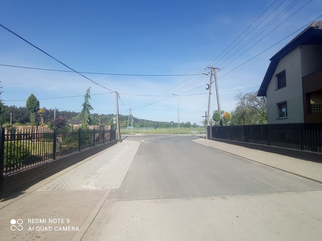 Zdjęcie przedstawia asfaltową drogę biegnącą przez wiejską okolicę. Po obu stronach drogi znajdują się ogrodzenia i domy. W tle widać pola i lasy. Na niebie jest bezchmurne, błękitne niebo.