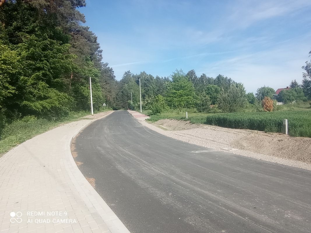 Zdjęcie przedstawia asfaltową drogę biegnącą przez zalesiony teren. Po lewej stronie drogi znajduje się chodnik z kostki brukowej. W tle widać drzewa i krzewy, a także kilka latarni ulicznych. Na niebie widoczne są lekkie chmury.