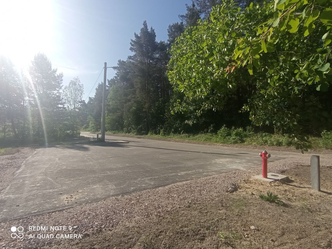 Zdjęcie przedstawia asfaltową drogę biegnącą przez zalesiony teren. Po prawej stronie drogi znajduje się czerwony hydrant przeciwpożarowy. W tle widać słup energetyczny oraz drzewa oświetlone słońcem. Na zdjęciu widoczny jest również cień rzucany przez drzewa.