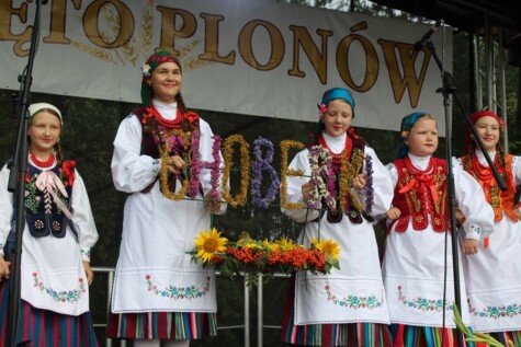 Grupa osób ubranych w tradycyjne stroje ludowe stoi na scenie podczas festiwalu. Trzymają ozdobny napis "CHOBERKI" wykonany z kwiatów. W tle widoczny jest baner z napisem "ŚWIĘTO PLONÓW".